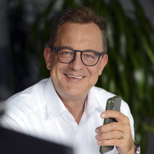 Ein lächelnder Mann mit Brille hält ein Smartphone in der Hand, umgeben von grünen Pflanzen im Hintergrund.